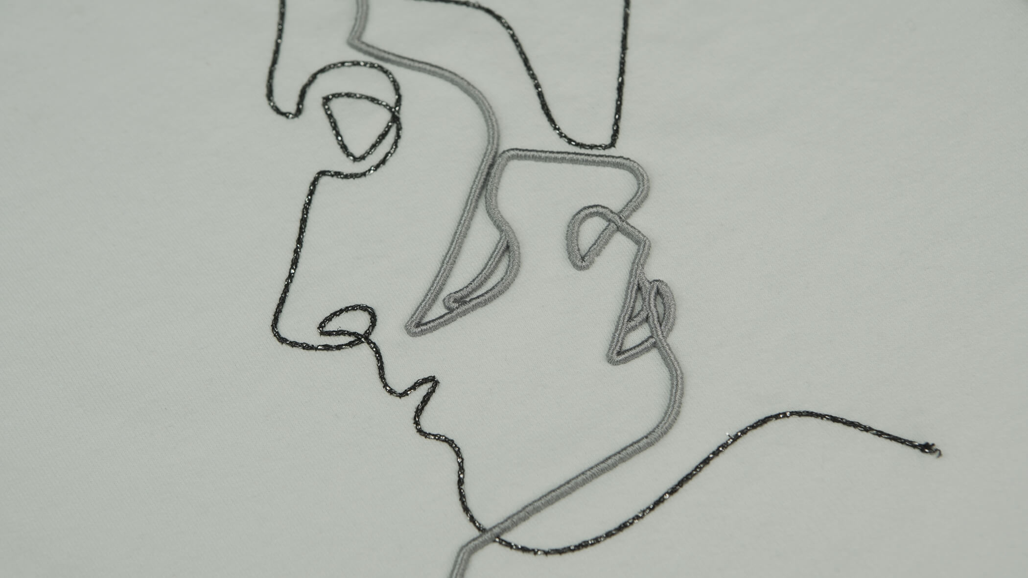 Dois rostos desenhados numa forma mais abstrata em cinzento e preto, com as técnicas de bordado com cordão e com correntes metálicas